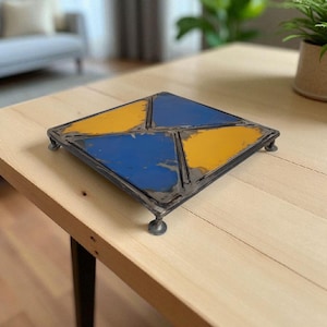 Könnte beinhalten: Ein quadratischer Metall-Topfuntersetzer mit einem Used-Look. Der Topfuntersetzer ist in vier Abschnitte unterteilt, die in Blau und Gelb lackiert sind. Er hat kleine, abgerundete Füße und einen dunklen Metallrahmen. Der Topfuntersetzer steht auf einem hellen Holztisch.