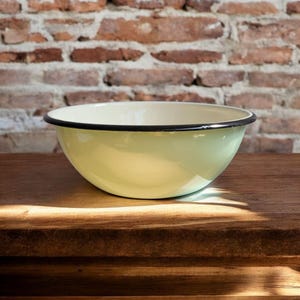 Könnte beinhalten: Eine hellgrüne Emaille-Schüssel mit schwarzem Rand. Die Schüssel steht auf einer Holzoberfläche vor einer Backsteinmauer. Die Schüssel ist rund und hat eine glatte, glänzende Oberfläche.