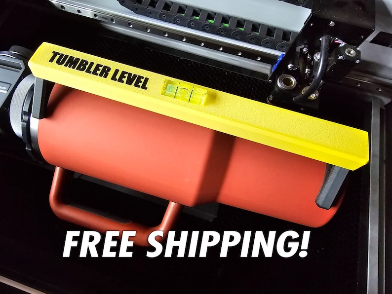 K&ouml;nnte beinhalten: Eine gelbe Wasserwaage mit der Aufschrift "Tumbler Level" ist an einem roten Becher befestigt. Der Becher wird in einer Lasergravurmaschine gehalten.