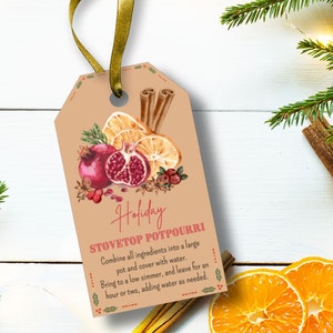 Könnte beinhalten: Ein beigefarbener Geschenkanhänger mit einer Aquarellillustration von Granatapfel, Orangenscheiben und Zimtstangen. Der Anhänger trägt die Aufschrift "Holiday STOVETOP POTPOURRI" mit Anweisungen zur Herstellung von Potpourri. Ein goldenes Band ist befestigt.