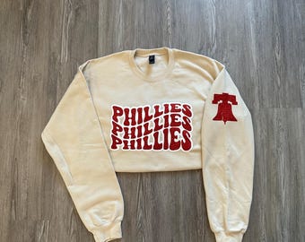May include: A cream-colored sweatshirt with a red "PHILLIES" logo on the front and a red Liberty Bell graphic on the right sleeve.