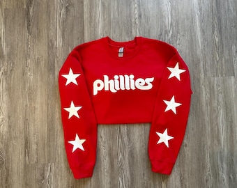 May include: Red cropped sweatshirt with white stars on the sleeves and the word "Phillies" in white on the front.
