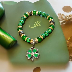 May include: A green and black beaded bracelet with a four-leaf clover charm. The bracelet is made with malachite and lava rock beads.