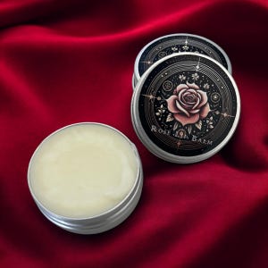 May include: A small silver tin with a white balm inside. The lid of the tin has a black background with a gold rose and a circle of gold stars and dots. The text "Rose Lip Balm" is printed in gold on the lid.