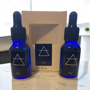 May include: Two cobalt blue glass bottles with black droppers and labels that read "Air" with a triangle symbol. A white box with the same symbol and text is behind the bottles. The bottles are 15 mL.