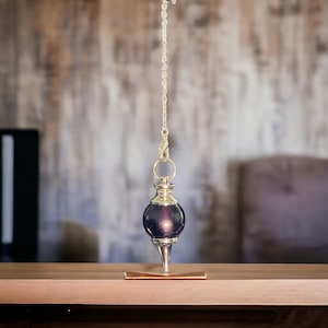 May include: A gold pendulum with a dark purple crystal bob hanging from a chain. The pendulum is resting on a wooden surface.