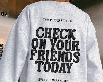 May include: A light gray sweatshirt with the text "THIS IS YOUR SIGN TO CHECK ON YOUR FRIENDS TODAY (EVEN THE HAPPY ONES)" printed in black on the back. The sweatshirt is a casual style.