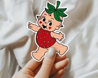 May include: A sticker of a cartoon baby wearing a strawberry costume. The baby has green hair and is smiling. The sticker is on a white background.