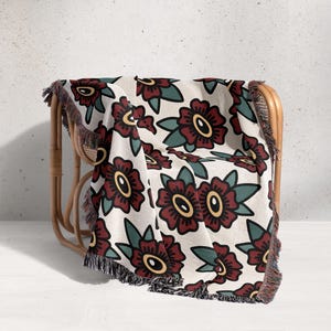 Puede incluir: Una manta de color crema con un estampado floral repetido en burdeos, amarillo y verde. La manta está sobre una silla de mimbre y tiene un ribete de flecos.