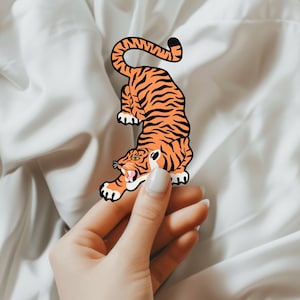 May include: A hand holding a tiger sticker with orange and black stripes. The sticker is on a white background.