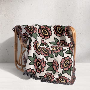 Puede incluir: Una manta con estampado floral sobre una silla de mimbre. La manta presenta un diseño repetido de flores rosas con centros amarillos y hojas verdes sobre un fondo crema. La manta tiene un borde con flecos.