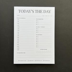 May include: A white notepad with the text "TODAY'S THE DAY" at the top. The notepad has sections for scheduling, top priorities, to-do list, water intake, and gratitude. It also includes checkboxes and a calendar.