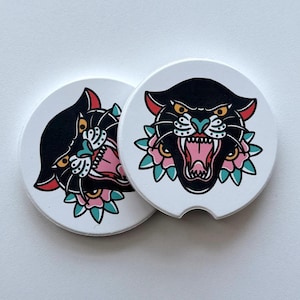 May include: Two white car coasters with a black panther design. The panther has red accents, yellow eyes, and a pink open mouth. The design includes pink and teal floral accents. The coasters are round and have a small notch.