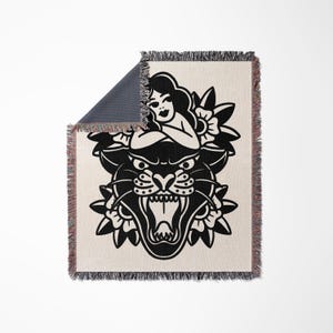 Puede incluir: Manta crema con un diseño de tatuaje de pantera negra. El diseño presenta una mujer, flores y una cabeza de pantera con la boca abierta. La manta tiene un borde con flecos y un reverso azul oscuro.