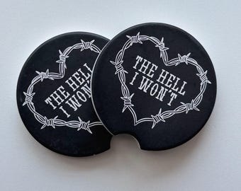 Puede incluir: Dos posavasos negros con texto blanco "THE HELL I WON'T" dentro de un diseño de alambre de púas en forma de corazón. Los posavasos son redondos y tienen una pequeña muesca.