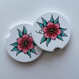 May include: Two white car coasters with a pink flower design. The flower has red outlines, a black center, and teal leaves. The coasters are round with a small notch on one side.