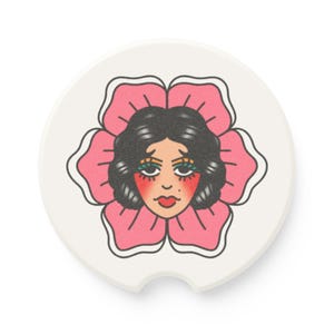 May include: A white car coaster featuring a floral design with a woman's face at the center. The woman has dark hair, red lips, and is surrounded by a pink flower with black outlines. The coaster has a curved notch for easy removal.