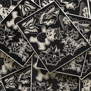 May include: Black and white temporary tattoo sheets featuring various traditional tattoo designs. The sheets include images of butterflies, flowers, a baby, and banners with the word "Lucky". The designs are in a classic tattoo style.