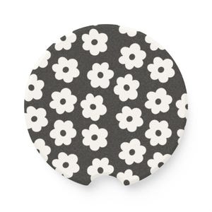 May include: A round car coaster with a black background and a repeating pattern of white daisy-like flowers. The coaster has a small notch for easy removal from the car's cup holder.