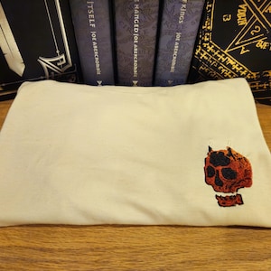 Puede incluir: Una bolsa de tela blanca con un diseño de calavera bordado en rojo y negro.