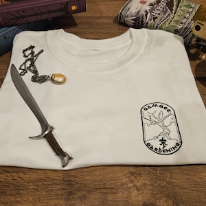 Op de afbeelding: Wit T-shirt met een zwart geborduurd logo met de tekst "Gamgee Gardening". Een zilveren dolk, een zwarte ketting met een hanger en een gouden ring liggen op het shirt. Een Funko Pop-figuur en een boek staan op de achtergrond.