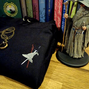 May include: A black hooded sweatshirt with an embroidered image of a wizard's hat, staff, and sword. The sweatshirt is folded and lying on a wooden surface. A figurine of a wizard with a staff and a long grey robe is in the background.