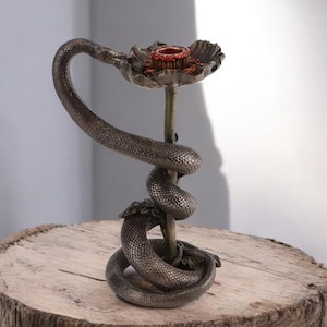 Può includere: Un portacandele color bronzo con un serpente avvolto attorno a uno stelo centrale. La parte superiore del supporto ha un design a forma di fiore con un centro rosso, progettato per contenere una candela. Il portacandele è posto su una superficie di legno.