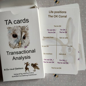 May include: A white card box with the text "TA cards" and "Transactional Analysis" with an owl illustration. The box is open, revealing cards with text and a rabbit illustration. The cards are labeled "Life positions The OK Corral".