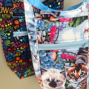 May include: Two colorful tote bags with cat designs. The bag on the left has a black background with colorful text and paw prints. The bag on the right has a blue background with a floral design and multiple cats. Both bags have a zippered pocket.