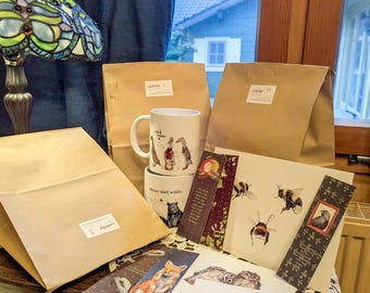 May include: A collection of nature-themed stationery and gifts. Includes a white mug with animal illustrations, brown paper bags, bookmarks, and art prints featuring a fox, hedgehog, and birds. The items are arranged on a wooden surface.