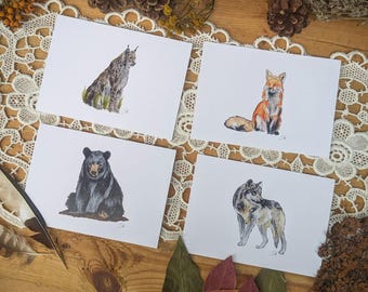 Può includere: Quattro biglietti bianchi con illustrazioni ad acquerello di animali della foresta: una lince, una volpe, un orso e un lupo. Le carte sono disposte su una superficie di legno con elementi decorativi.