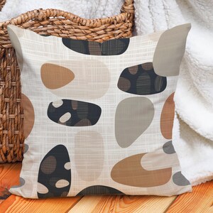 May include: Decorative throw pillow with an abstract pattern of organic shapes in shades of brown, beige, and black. The pillow has a textured, off-white background and is placed near a woven basket and a white blanket.