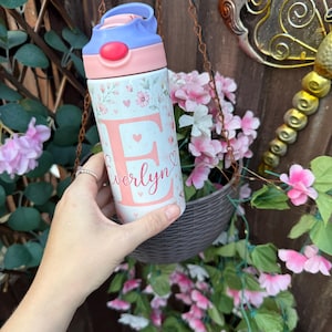 May include: A white water bottle featuring a floral pattern with small pink flowers and hearts. The letter E is printed in a large pink font with the name Everlyn written in a cursive script across it. The bottle has a light purple and pink lid.
