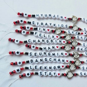 May include: A set of 10 beaded bracelets with the names of the members of the K-pop group Stray Kids, including the words "Stray Kids", "SKZ", "In", "Felix", "Changbin", "Han", "Seungmin", "Bangchan", "Lee Know", and "Stay". Each bracelet features a red heart charm and a silver star charm.