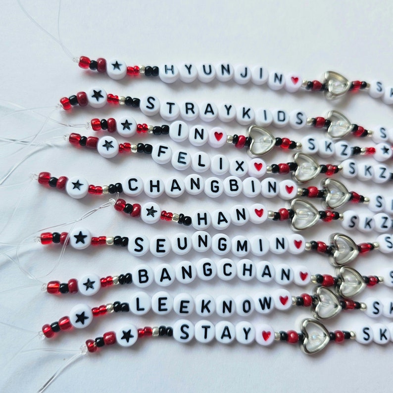 May include: A set of 10 beaded bracelets with the names of the members of the K-pop group Stray Kids, including the words "Stray Kids", "SKZ", "In", "Felix", "Changbin", "Han", "Seungmin", "Bangchan", "Lee Know", and "Stay". Each bracelet features a red heart charm and a silver star charm.