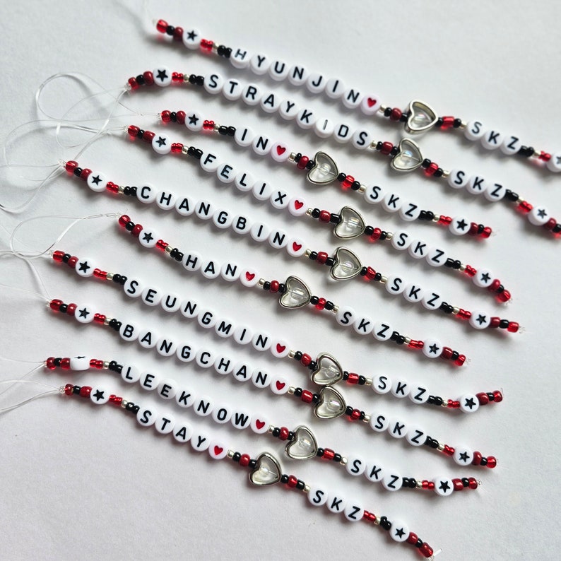 May include: A set of 10 beaded bracelets with white beads that spell out the names of the members of the K-pop group Stray Kids. Each bracelet has a silver heart charm and red and black beads.