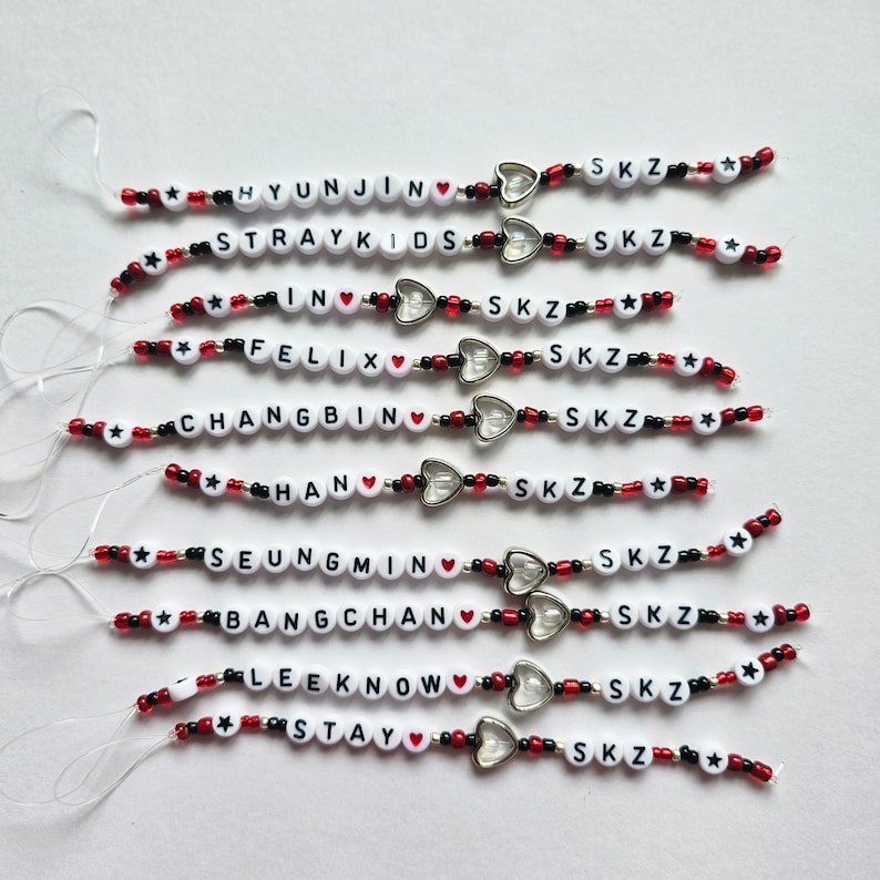 May include: A set of ten phone charms featuring the names of the members of the K-pop group Stray Kids, each with a red heart and the letters "SKZ" in black beads.