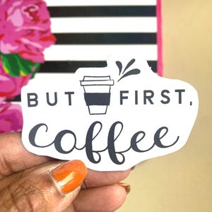 Puede incluir: Una pegatina blanca con el texto "BUT FIRST, coffee" en escritura negra, con un gr&aacute;fico de taza de caf&eacute;. La pegatina se sostiene frente a un fondo con rayas blancas y negras y elementos florales rosas.