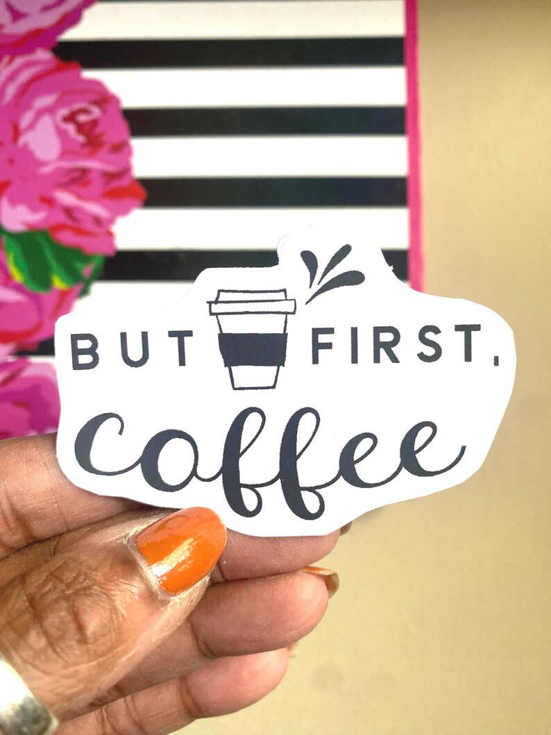 Puede incluir: Una pegatina blanca con el texto "BUT FIRST, coffee" en escritura negra, con un gr&aacute;fico de taza de caf&eacute;. La pegatina se sostiene frente a un fondo con rayas blancas y negras y elementos florales rosas.