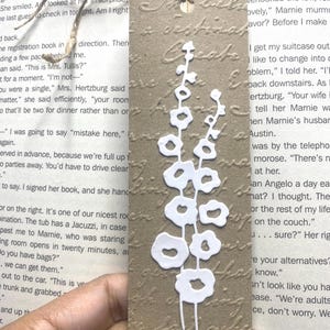 May include: A brown paper bookmark with a white floral design. The bookmark has a textured surface and a string for hanging. The floral design is a stylized stem with several flower shapes.
