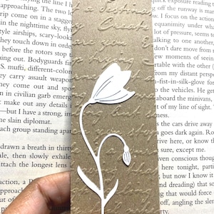May include: A brown paper bookmark with a white floral design. The bookmark has a textured surface with embossed text and a hole at the top for a string. The floral design is a stylized flower with a curved stem.