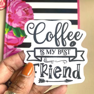 Puede incluir: Una pegatina blanca con el texto "Coffee is my best friend" en escritura negra y letras de bloque. La pegatina se sostiene frente a un cuaderno con un dise&ntilde;o floral y rayas blancas y negras.
