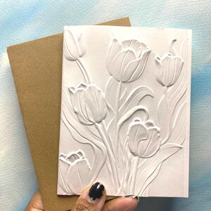 May include: A white embossed greeting card featuring a floral tulip design, held with a brown envelope. The card has a textured surface with raised tulip flowers and leaves. The background is a light blue and white watercolor effect.