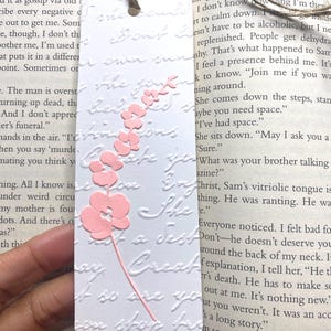 May include: A white bookmark with embossed text and a pink floral design. The bookmark has a jute twine for hanging and is resting on the pages of a book. The floral design features a stem and several flower blossoms.