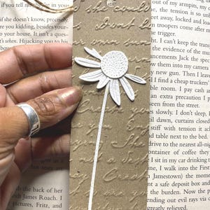 May include: A handmade bookmark with a white daisy design. The bookmark is rectangular with rounded corners and a textured, brown paper appearance. A white daisy with a long stem is affixed to the bookmark, and a twine loop is at the top.
