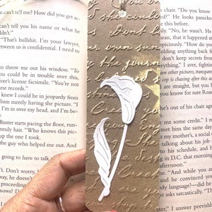 May include: A handmade bookmark with a calla lily design. The bookmark is a rectangular shape with a textured, brown background and a white calla lily and leaf design. A twine is attached at the top.