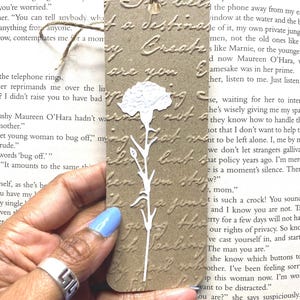 May include: A brown bookmark with embossed script and a white floral cut-out design. The bookmark is rectangular with a jute twine attached. The text on the bookmark reads "...a destination...Create...are you...but..."