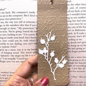 May include: A brown paper bookmark with a white floral design and a jute string. The bookmark has embossed text and a die-cut botanical element. A simple, elegant accessory for book lovers.