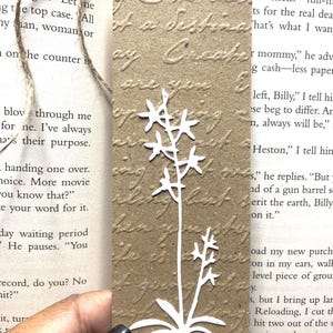 May include: A rectangular brown paper bookmark featuring an embossed cursive script texture across the surface. A white die-cut floral silhouette with a central stem and small star-shaped blossoms is centered on the bookmark, which has a jute twine tie.