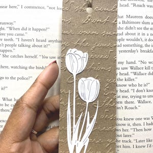 May include: A brown paper bookmark with a white tulip design. The bookmark has a textured surface and a hole at the top with a twine string. The background is a book page.